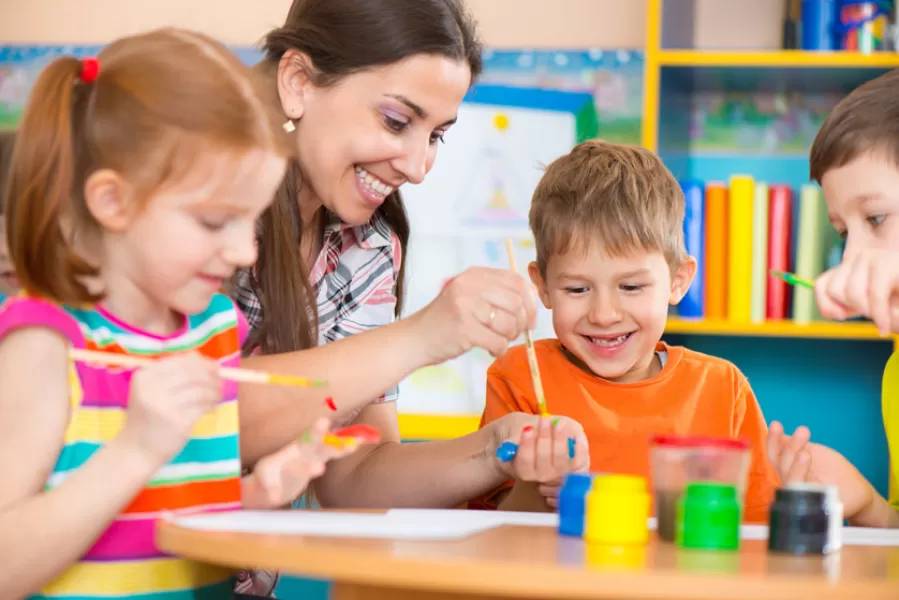 A teacher paints with kids at a Preschool arts and crafts table while smiling